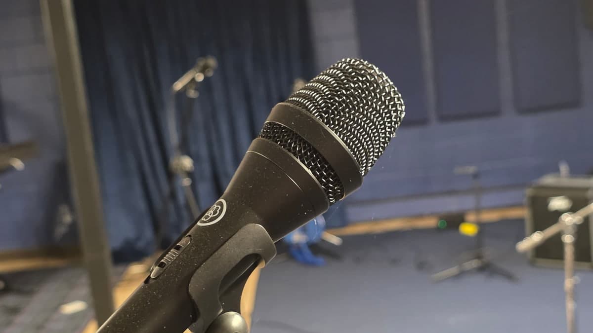 An AKG microphone on a boom stand in close focus, with drapes, empty stands and flight cases blurred behind it.