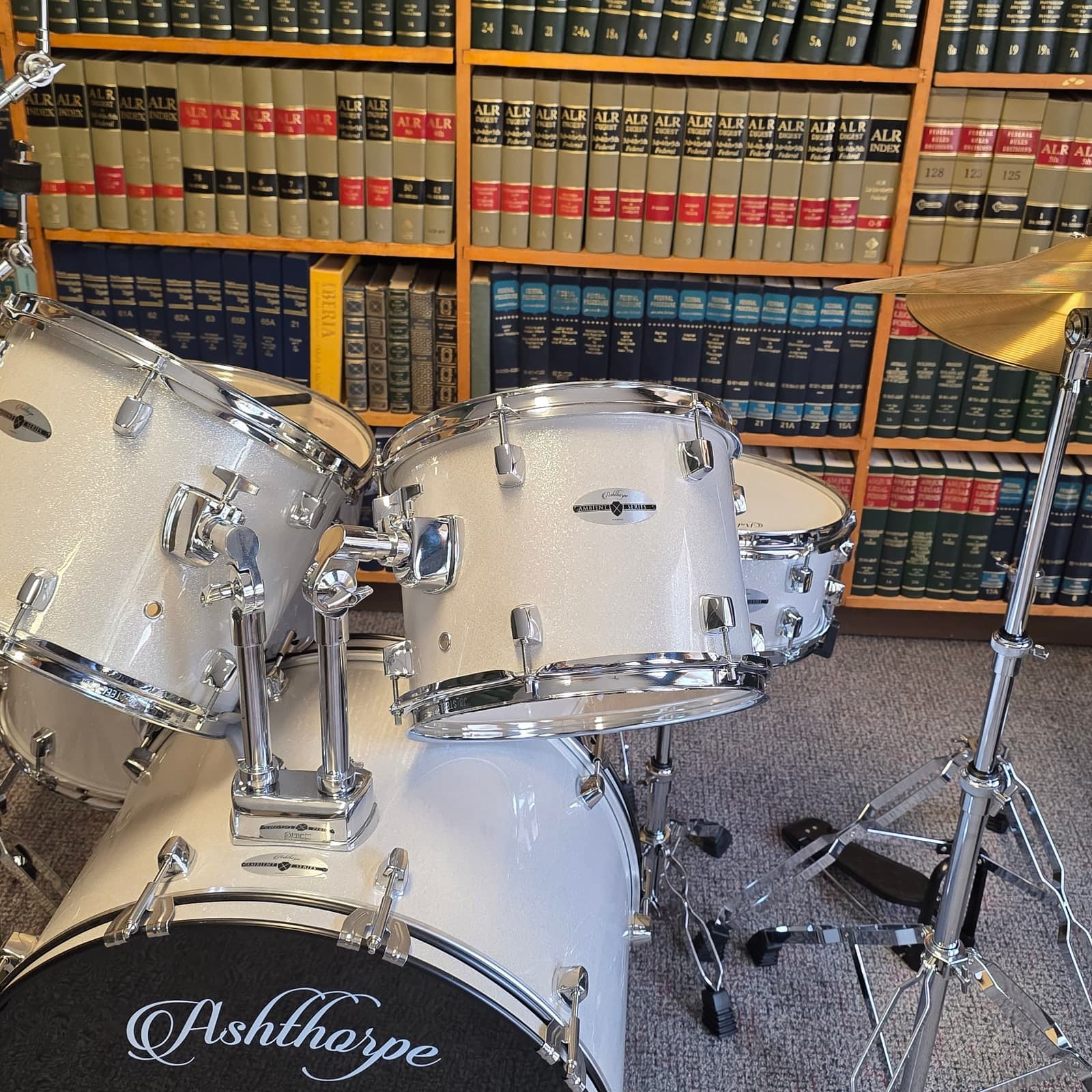 A white Ashthorpe drum kit and cymbal set up in front of floor-to-ceiling library shelving.