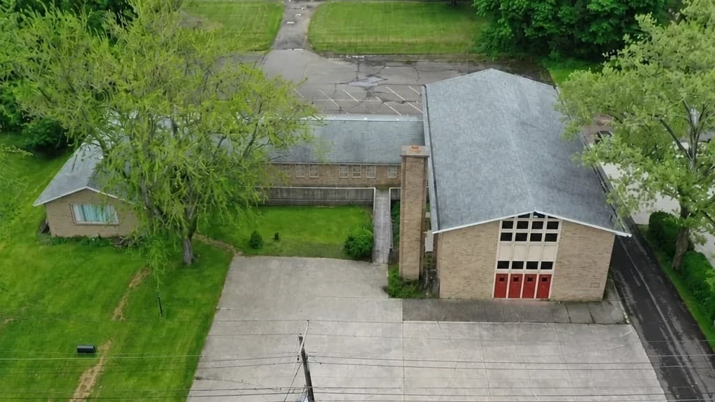 Power Chord Studios from the air: a brick hall with a steep gabled roof and a tall window wall above four red doors, a lower wing running back from it, and a concrete lot down to the street.