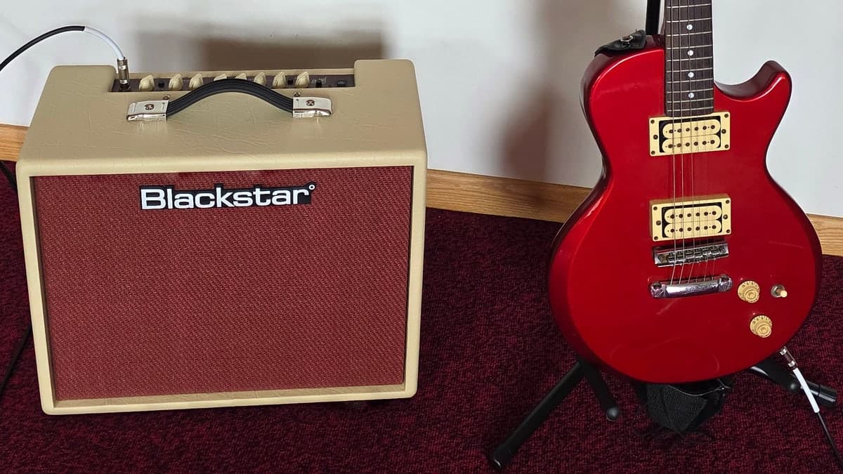 A red Lotus solid-body electric guitar on a stand beside a cream and oxblood Blackstar combo amplifier, on red carpet against a white wall.